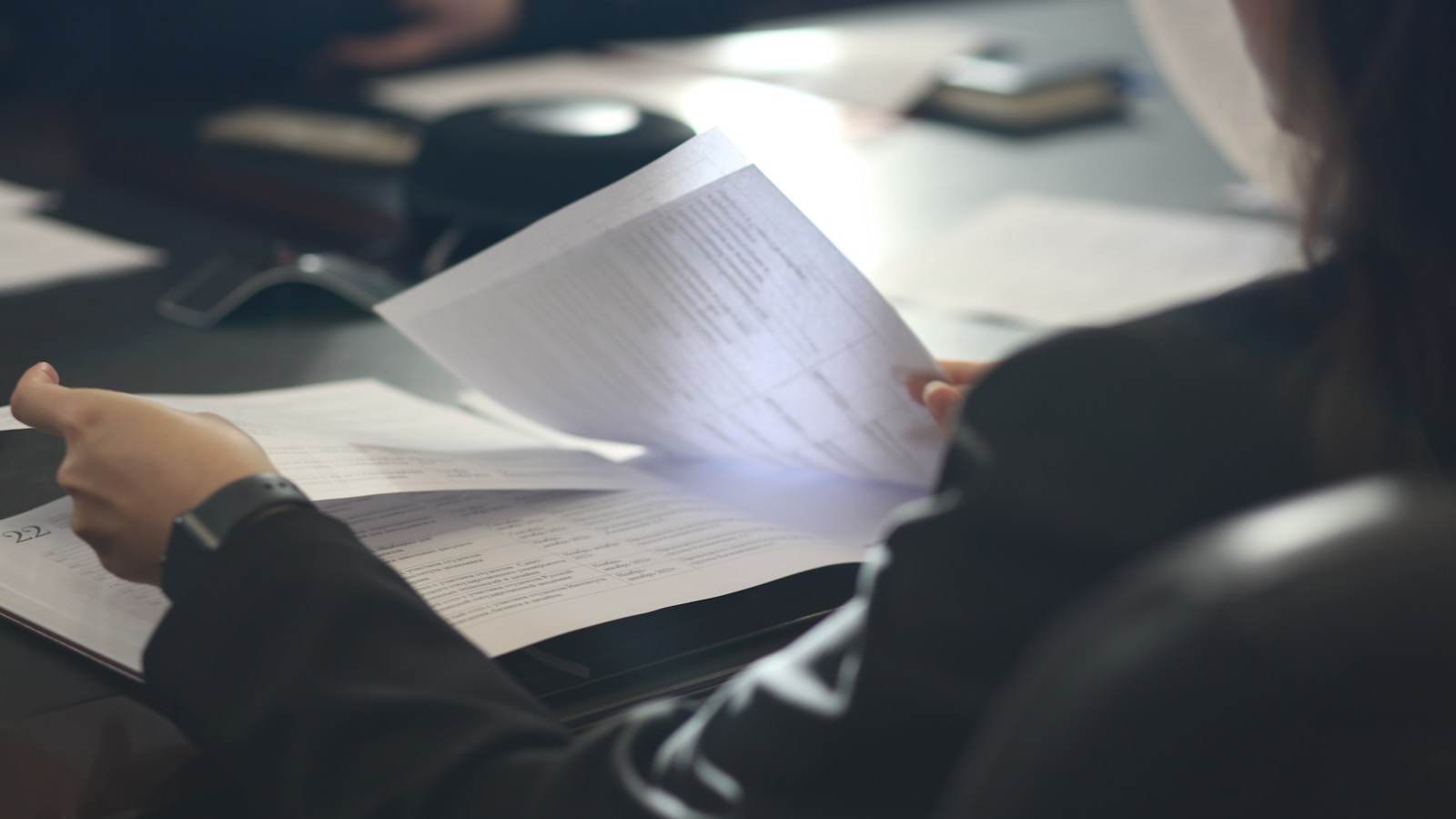 Une personne relit attentivement un document imprimé posé sur un bureau, page soulevée entre les doigts